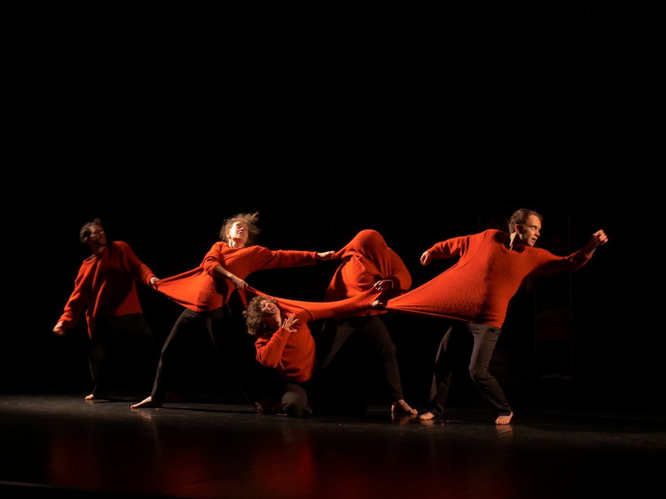 Cinq danseurs en pulls rouges s'entremêlent et s'étirent dans un mouvement collectif sur scène.